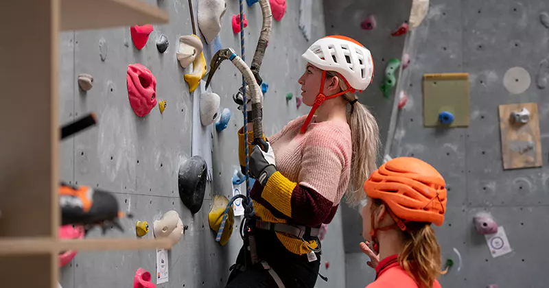 Twee mensen met helmen en klimgordels zijn aan het indoor klimmen. De ene beklimt de wand met ijsbijlen, terwijl de andere vlakbij staat toe te kijken en aanwijzingen geeft. Op de wand zijn kleurrijke klimgrepen te zien.